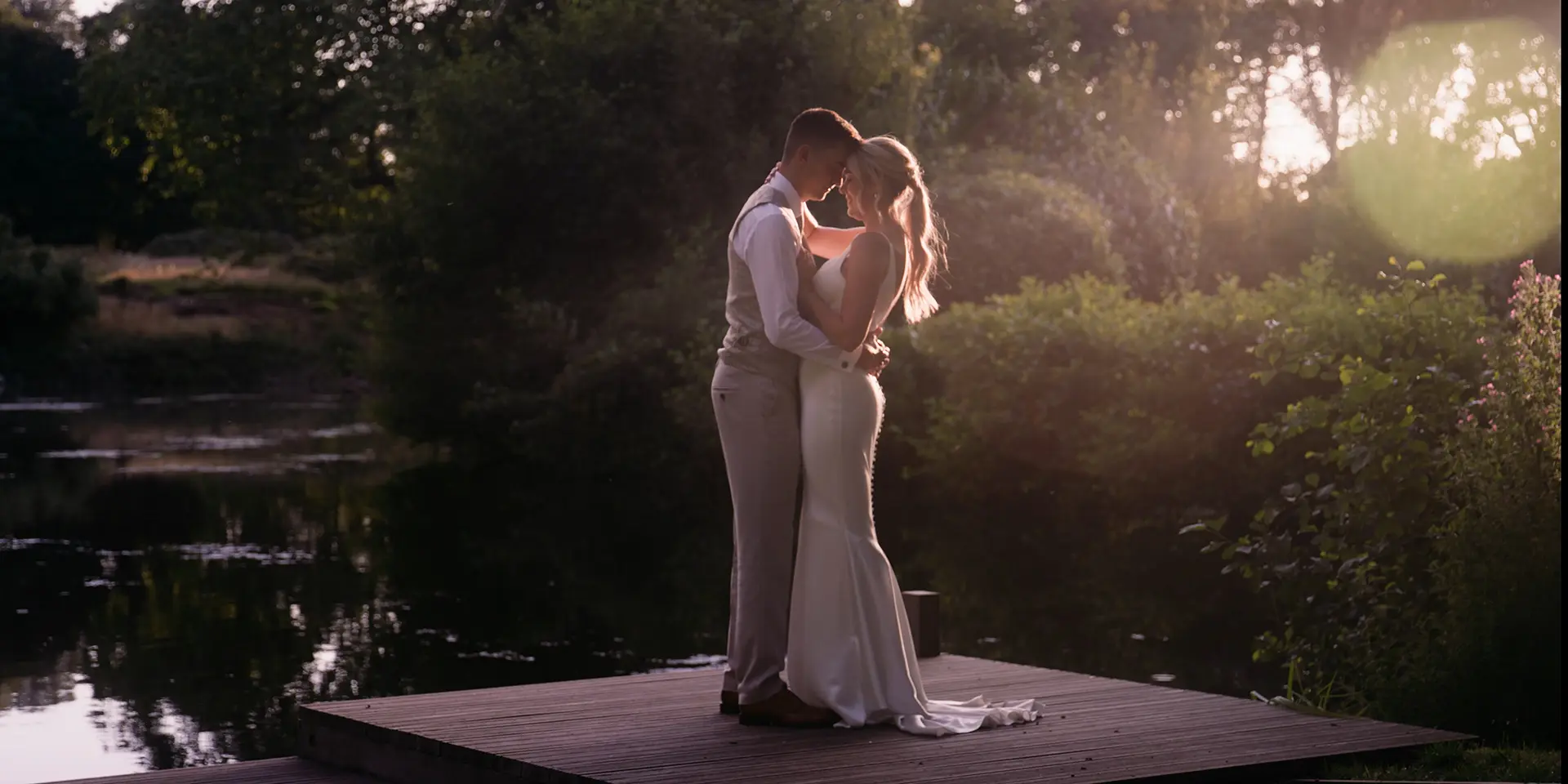 Still frame from a wedding video featuring a bride and groom kissing at sunset near a lake.